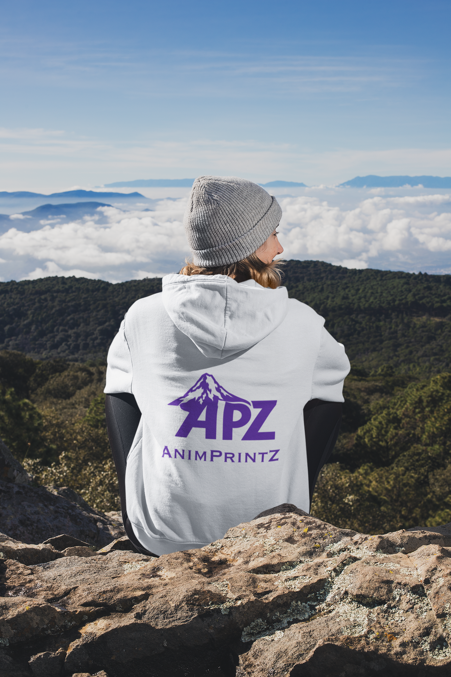 Woman with white hooded sweatshirt sitting on a rocky surface overlooking a mountain and forest landscape. The back of the hoodie shows the AnimPrintZ logo - APZ with a mountain and AnimPrintZ below that.