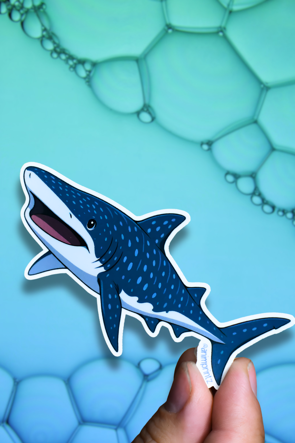 A die-cut sticker of a cartoon whale shark being held by a person's fingers against a blue background with bubble graphics.